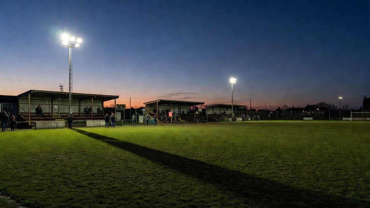Small floodlit lower-league football ground at dusk